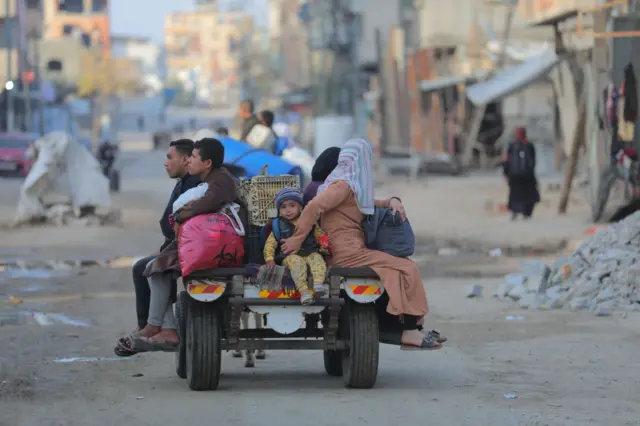 A man, boy, young child and two woman sit on the back of a vehicle. The woman holds the child so he is secure. They have bags, and drive amongst rubble and dirt, looking downcast