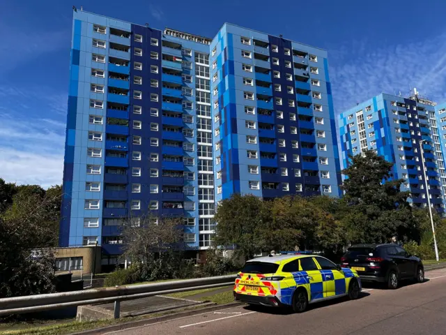 A police car parked in front of three large, blue blocks of flats on a sunny day.