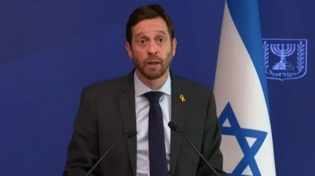 A man in a suit jacket standing behind two small microphones. He stands in front of a blue background, and an Israeli flag. He speaks into the microphones