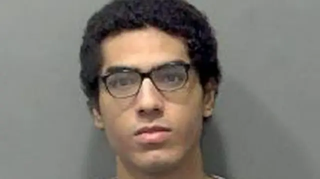 Custody mugshot of Nicholas Prosper. He has curly black hair and is wearing black glasses. He has a serious expression on his face.