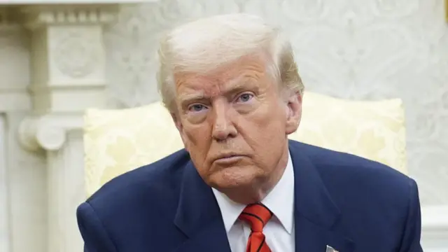 Donald Trump, wearing a blue suit with red tie, is photographed from the shoulders up. He is sitting on an ornate cream chair in front of the white wallpaper of the oval office, looking directly into the camera, his head tilted to the left, with a neutral expression