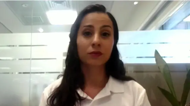 Farsakh, who has brown hair, wears a white polo shirt and sits in front of frosted glass with the leaves of a plant visible just behind her