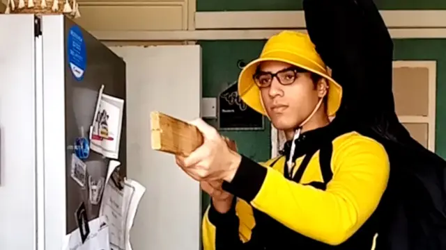 Nicholas Prosper wears a black and yellow hoodie as he holds a plank of wood. He is aiming the wood as if it is a gun and he is standing in a kitchen, next to a fridge.