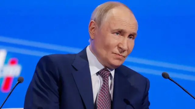 Putin looks ahead and sits behind a microphone on stage wearing a suit.