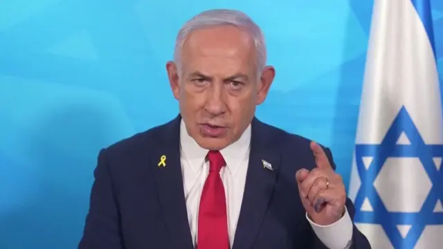 Netanyahu wears a navy suit and red tie as he speaks. He stands in front of an Israeli flag set against a blue background