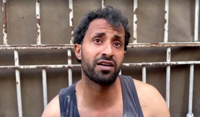 A man with curly brown hair sittin down in front of a metal gate. He speaks and looks slightly above the camera. He wears a black vest and has some dust on it