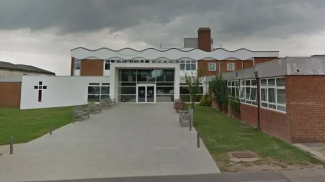 The entrance to Cardinal Newman school. It has a Christian cross on a wall, with a glass entrance to the building. There is a paved walkway leading to the doors and this is lined with benches.