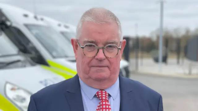 John Tizard in a blue suit and shirt with a red and white tie. He is wearing glasses and standing in front of a fleet of police vans