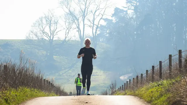 Jamie running over a hill in the English countrside with an official in hi-vis behind him