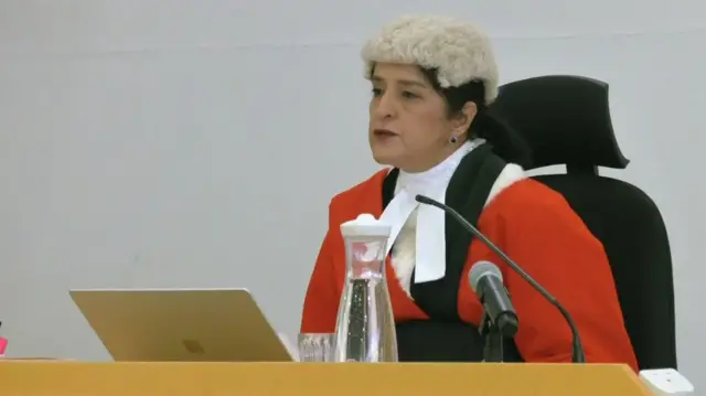 Mrs Justice Cheema-Grubb, wearing a wig, red gown and sitting in a chair behind a wooden desk. She is reading her sentencing remarks from a laptop.