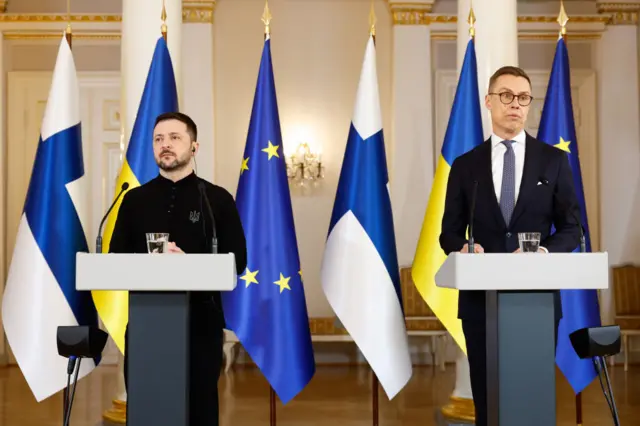 Zelensky on the left, Stubb on the right in front of their countries respective flags. They both stand at lecterns.