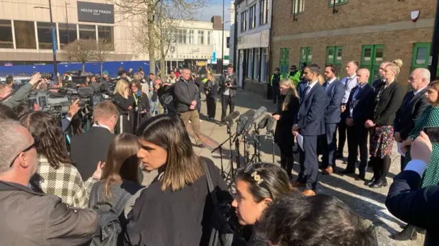 A large crowd of reporters filming and holding microphones towards a number of police staff, all wearing suits or dresses.