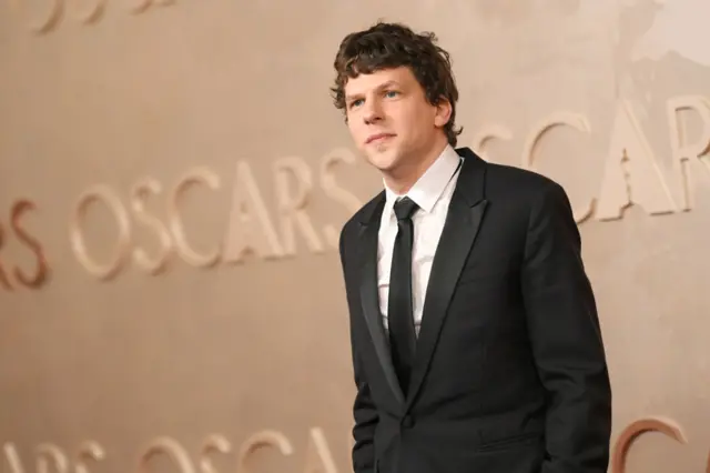 Jesse Eisenberg wearing a black suit and tie with white shirt smiles on the red carpet