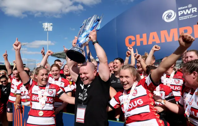 Sean Lynn holds the Premiership Women's Rugby trophy in the air surrounded by the players after winning the 2024 final