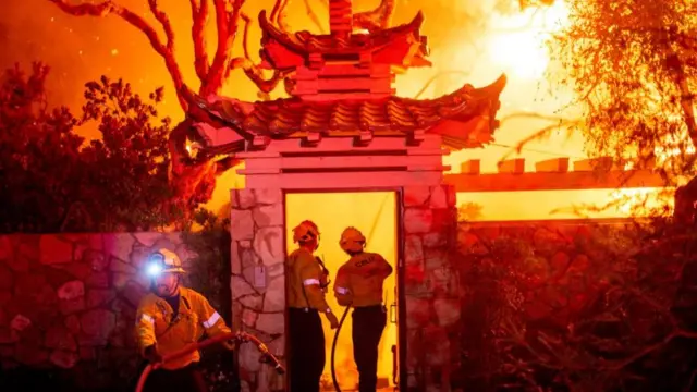 Three firefighters battle a blazing fire behind the entrance of a house's gates