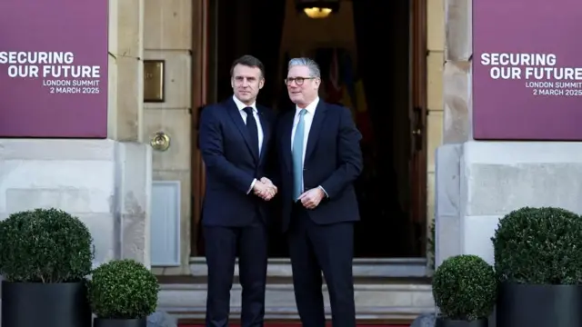 Starmer greets Macron, shaking hands outside large building.