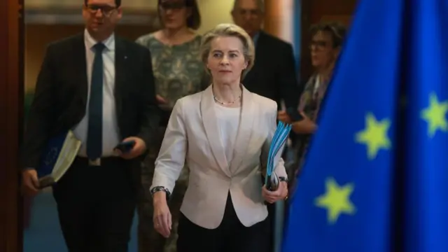 European Commission President Ursula von der Leyen walks ahead, holding folders, wearing a blazer. Several people are gathered behind her.