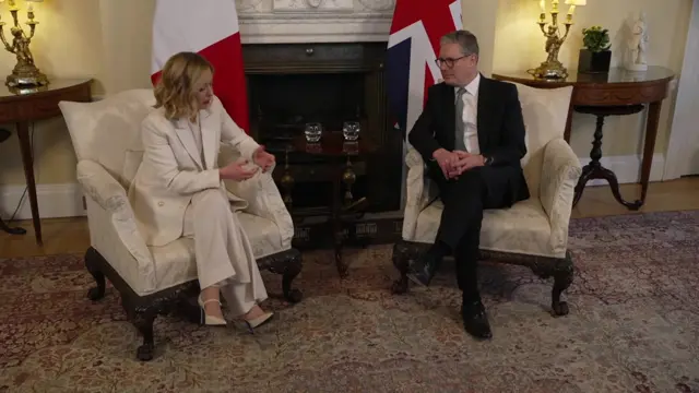 Meloni and Starmer sit by a fireplace for a conversation. They're respective flags, Italian and British, are draped behind them