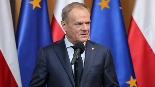 Poland's PM Donald Tusk wears a suit in front of flags of the EU and Poland. He speaks behind a microphone.