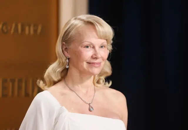 Pamela Anderson wearing a white one shoulder dress and pendant necklace