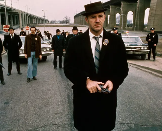 Policeman Gene Hackman is shown in The French Connection still walking toward the camera. He is holding a gun in his hands with policemen and police vehicles in the background. still walking toward the camera. He is holding a gun in his hands with policemen and police vehicles in the background.