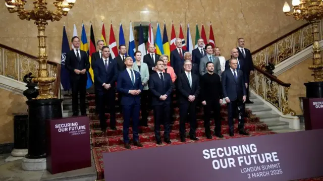 British Prime Minister Keir Starmer and Ukrainian President Zelensky pose for a family photo with fellow leaders. Flags are lined up in the background