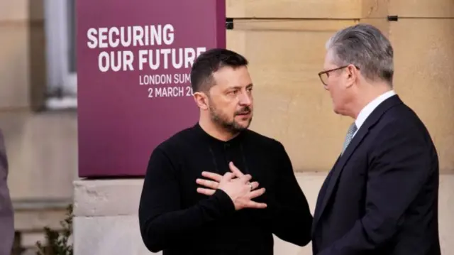 Volodymyr Zelensky (L) with his hands crossed on his chest as he speaks to Keir Starmer (R). Zelensky is in a black shirt, Starmer in a dark suit with azure tie and white shirt, they're standing outside Lancaster House