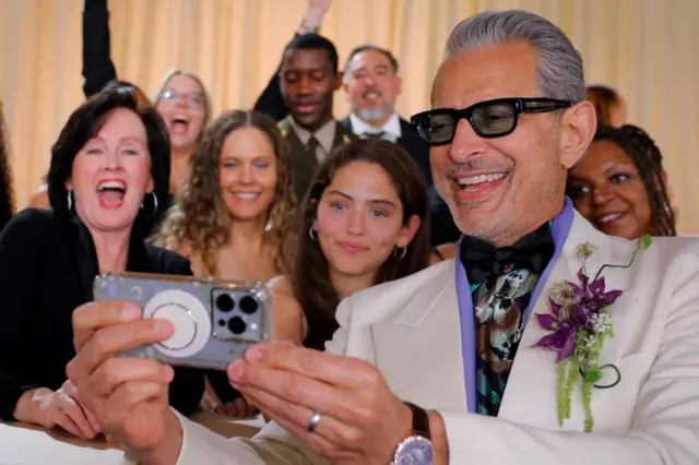 Jeff Goldblum takes a selfie with fans on the red carpet, wearing a cream blazer with purple flours on a brooch