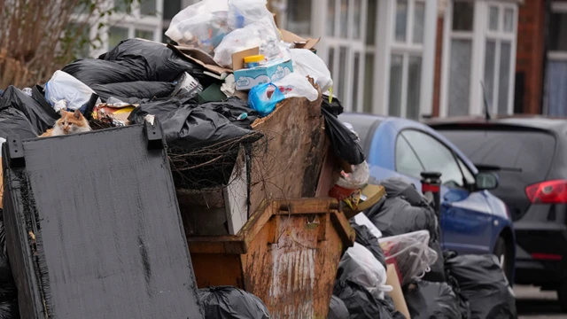 Rubbish bags overflowing from a yellow skip next to fly-tipping, with cars in the background.