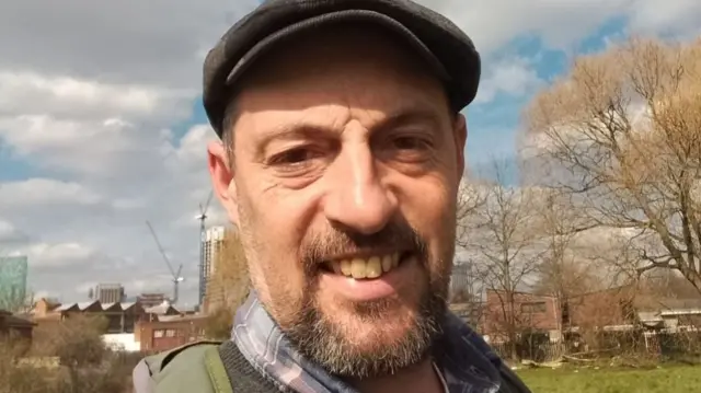 A man with a flat cap and a dark beard standing in an area with trees and industrial buildings behind him.