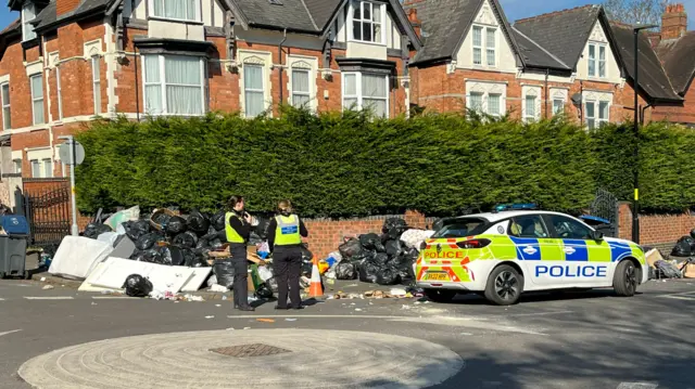 Police officers stand next to a police car with large piles of rubbish almost as tall as the officers behind them