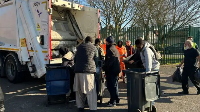 Male residents pushing bins towards a refuse lorry. Around six men are seen in the picture, with a further three refuse workers in bright orange standing in the background. The lorry is stationary on the side of the street and rubbish has been dropped around it on the floor.