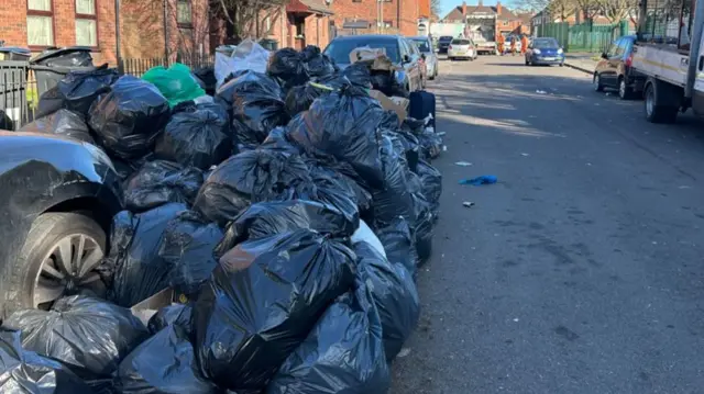 Bin bags stacked to the side of a residential street next to parked cars, blocking one in.