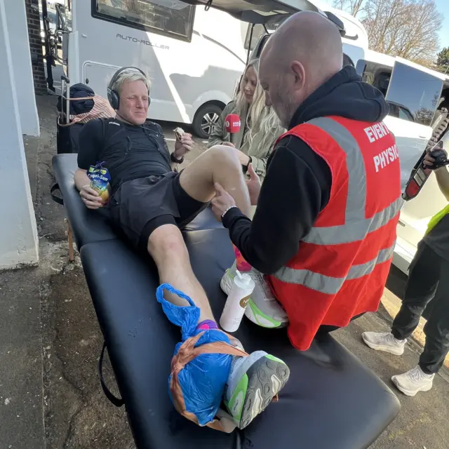 Jamie Laing lay down receiving physio treatment on his knee while holding a drink and a snack