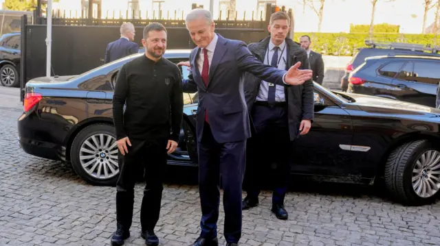 Clad in black, Ukrainian President Volodymyr Zelenskiy meets Norwegian Prime Minister Jonas Gahr Stoere, who wears a navy suit and red tie with one arm outstretched and another behind Zelensky's back.