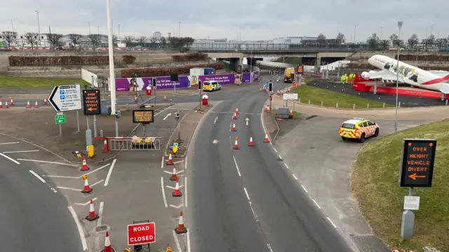 The road coming into heathrow airport is deserted of cars. road works signs and closure signs can be seen.