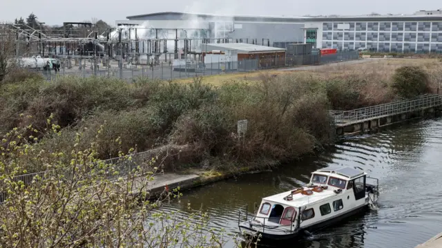 A boat on a river in front of smoke rising from the area of an electrical substation