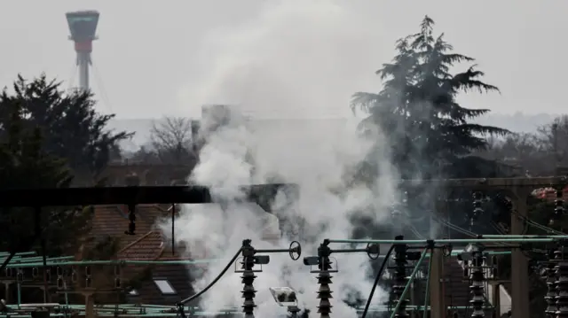 Smoke rises from the area of an electrical substation, with a control tower from the Heathrow International Airport on the background