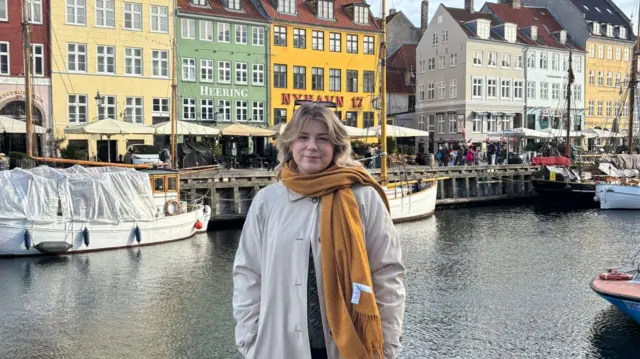 Annabelle (young woman with long blond hair with a yellow scarf around her neck) poses for pictures in front of coloured houses in Nyhavn port in Copenhagen in a beige trench coat. There's three wooden boats moored behind her