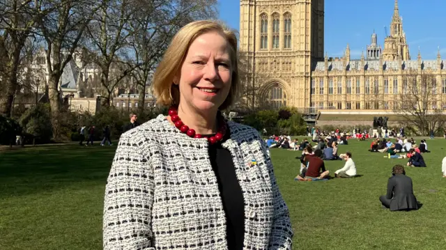 Ruth Cadbury MP posing on the green outside the Houses of Parliament (visible in the background). On the patch of grass behind her are several people sitting down in the sunshine