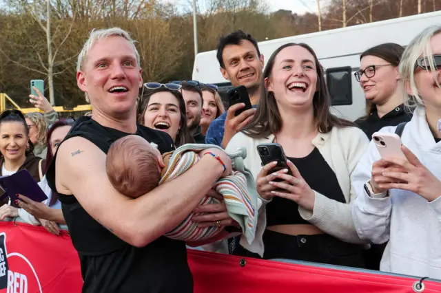 Jamie smiling and holding a baby in front of a group of people smiling