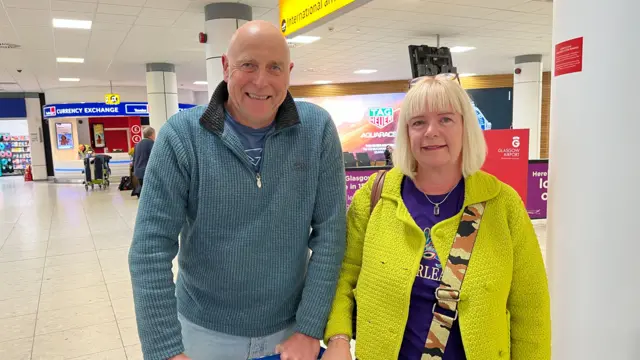 Simon (L) and Vanessa (R) pose for picture inside Glasgow airport. Simon is in a blue grey sweathshirt and jeans while Vanessa is in a yellow jacket with a blue shirt undereneath