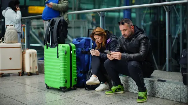 two passengers surrounded by luggage sit on a ledge at heathrow airport. they're using their mobile phones.