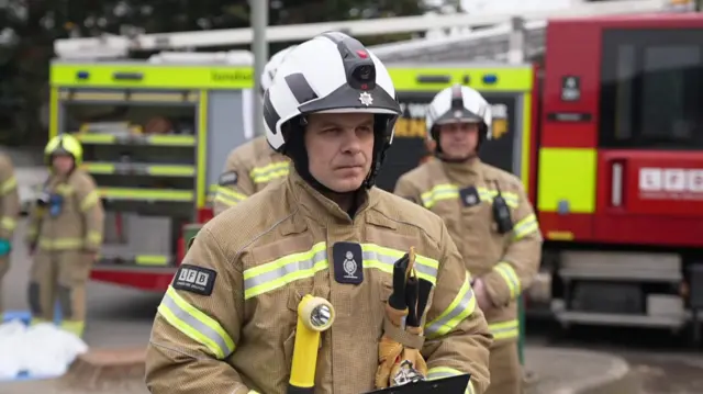 A firefighter wearing his uniform and helmet stands in front of other firefighters and a fire engine and addresses the media behind the camera