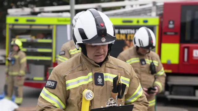 A firefighter wearing his uniform and helmet stands in front of other firefighters and a fire engine and addresses the media behind the camera