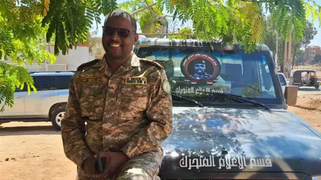 A man in military uniform sits on the bonnet of a jeep.