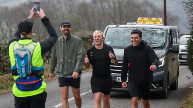 Three men running along a country lane with a taxi behind him.