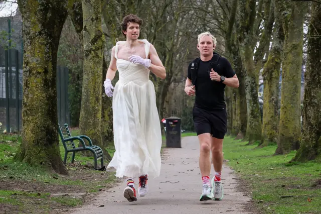 Two men running in a park. One is wearing black shorts and t-shirt, and the other is an a wedding dress.