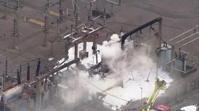 Emergency workers attend the scene at the substation, as viewed from the air. It is producing some kind of white smoke or steam, and appears to be covered in foam