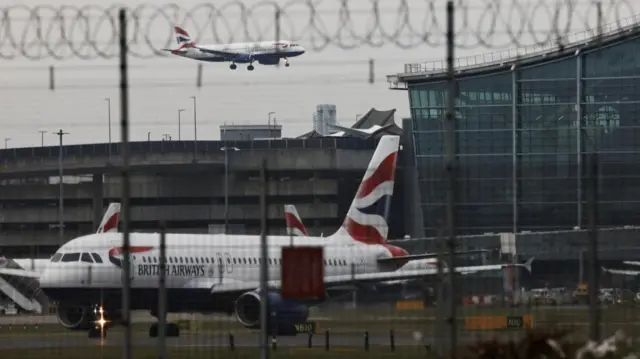 A British Airways plane in the sky as another can be seen on the ground. Image has been taken through a metal fence